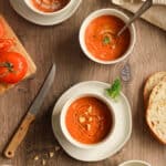 Gluten free tomato soup in three white bowls on a wood surface with a knife and tomato on a cutting board next to it and bread as well as a napkin