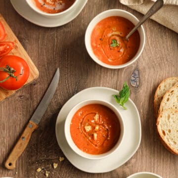 Gluten free tomato soup in three white bowls on a wood surface with a knife and tomato on a cutting board next to it and bread as well as a napkin