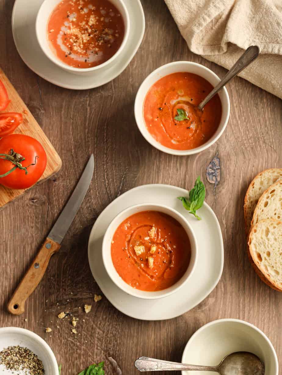 Gluten free tomato soup in three white bowls on a wood surface with a knife and tomato on a cutting board next to it and bread as well as a napkin.