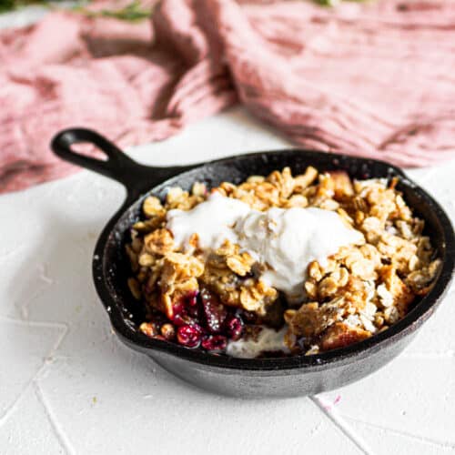 Skillet Apple Crisp in maroon towel and flowers in background. Half of crisp is mixing to show inside with blueberries and apples