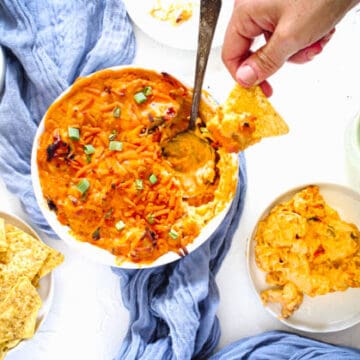 Vegan Buffalo Dip in a casserole dish with a chip with the dip of top close by the bowl. the dip is on a blue towel