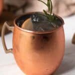 apple moscow mule in a mule glass with rosemary garnish on a white marble table