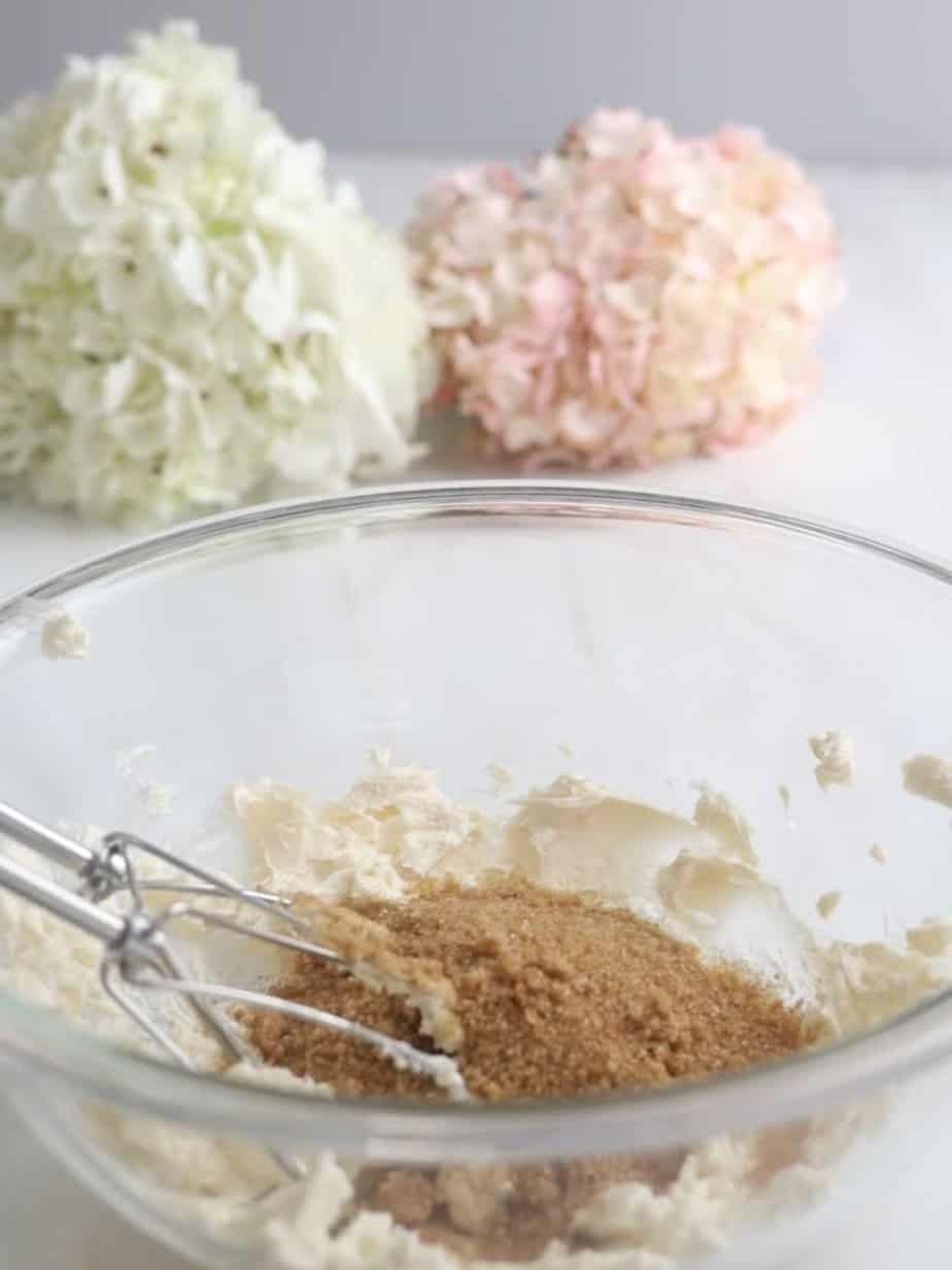 Sugar and butter mixing in a mixing bowl with flowers in the background and electric mixer in bowl