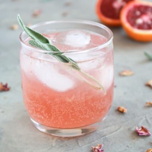 Pink gin cocktail in a glass with sage in the glass and rose petals surrounding it on a gray surface