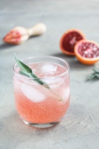 Sparkling Blood Orange Gin Cocktail in aglass with sage in the glass and ice. There is a blood orange and juicer in the back on a grey surface