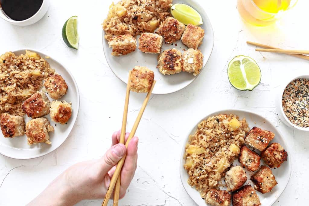 Coconut crusted tofu with sesame seeds plated on three plates with pineapple rice next to it. There are also chopsticks with one tofu piece being pulled away and clear beverages within the scene