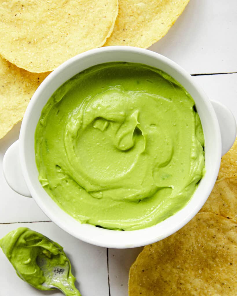 Creamy avocado dip in a white bowl on a textile surface with tortillas