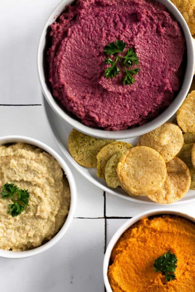 three bowls of carrot hummus in multiple colors (purple yellow and orange) with chips around it on a white surface