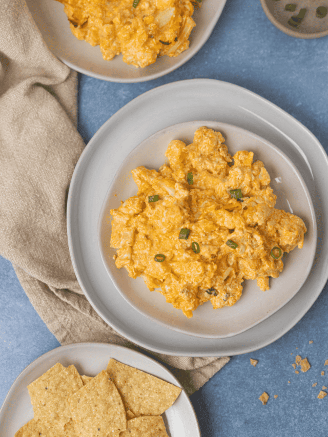 Vegan buffalo chicken dip on a white plate with green chives on top and tortilla chips next to it, as well as extra dip on another white plate next to it and a bowl of chives on top of a linen napkin