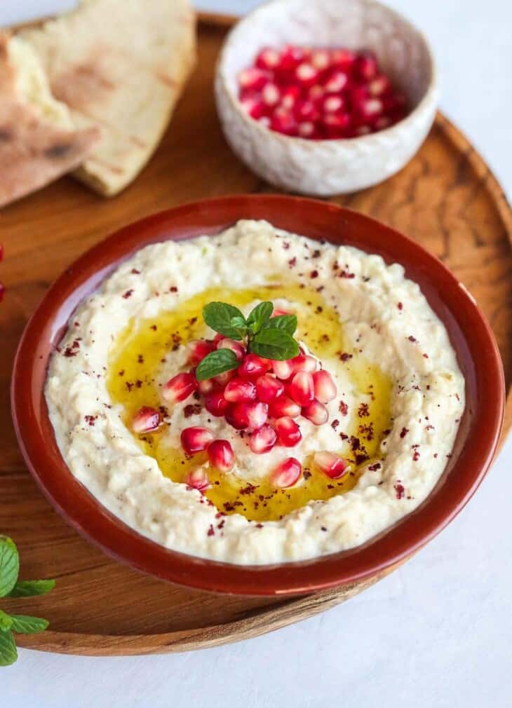 Mutabbal in a bowl with olive oil and pomegranate seeds on top. thed red bowl is on a wood plate.