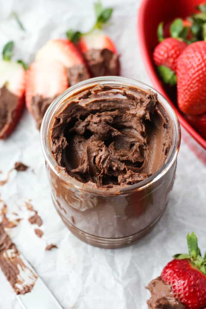 Nut Free chocolate spread on a glass jar on parchment paper with strawberries dipped in chocolate next to it and a knife with chocolate