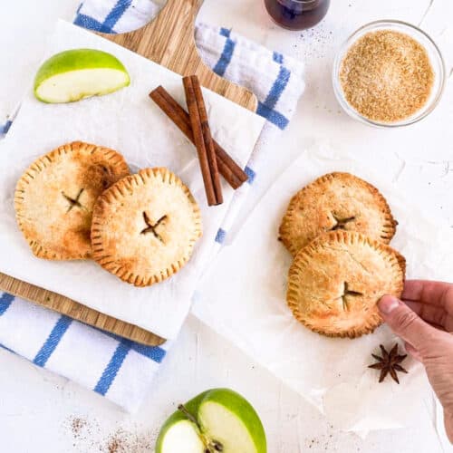 Vegan hand pie on a cutting board with two other hand pies off cutting board and apples surronding it. There are also cinnamon sticks next to it and a bowl of sugar