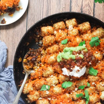 vegan tater tot casserole in a cast iron skillet topped with guacamole, salsa, and cilantro