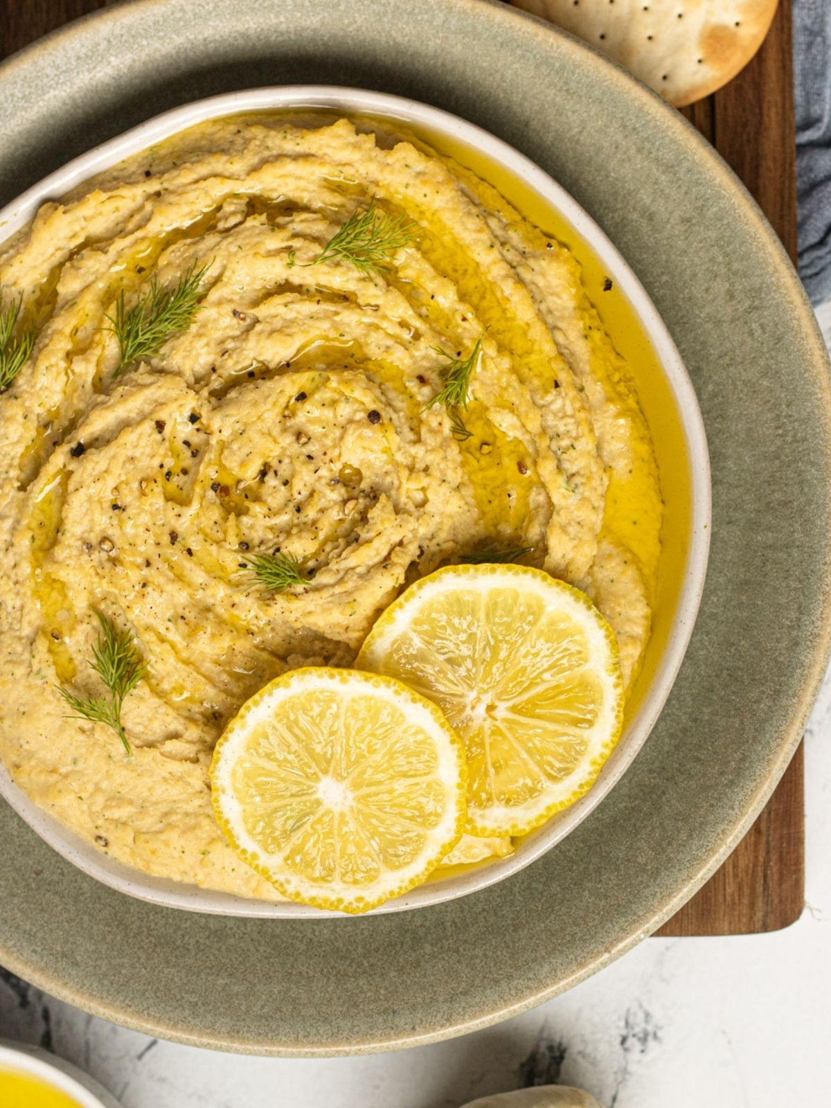 Dill Hummus on a white plate, on top of a cutting board. The hummus is topped with lemon, fresh dill, olive oil and salt + pepper