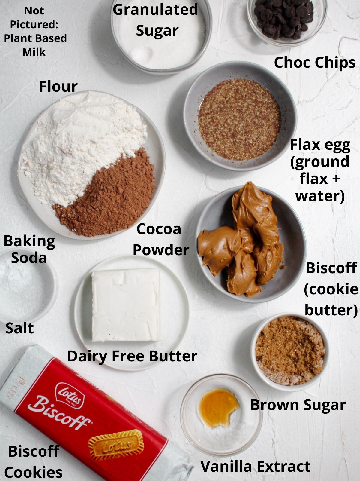 All ingredients for vegan biscoff brownies in bowls on a white surface. Each ingredient is labeled with black text.