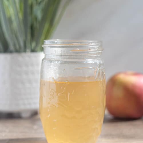 apple simple syrup in a mason jar on a wood table with a plant and apple behind it