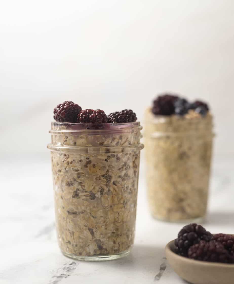 Overnight oats with frozen blackberries on top in a mason jar on a marble surface and a bowl of frozen fruit in the front