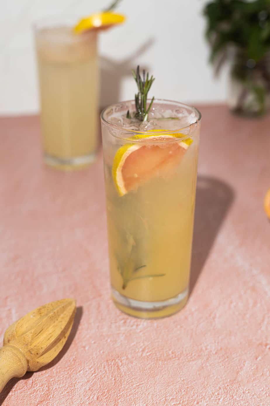 Two grapefruit rosemary vodka seltzer in highball glasses with rosemary garnish on a pink surface at a 90 degree angel.