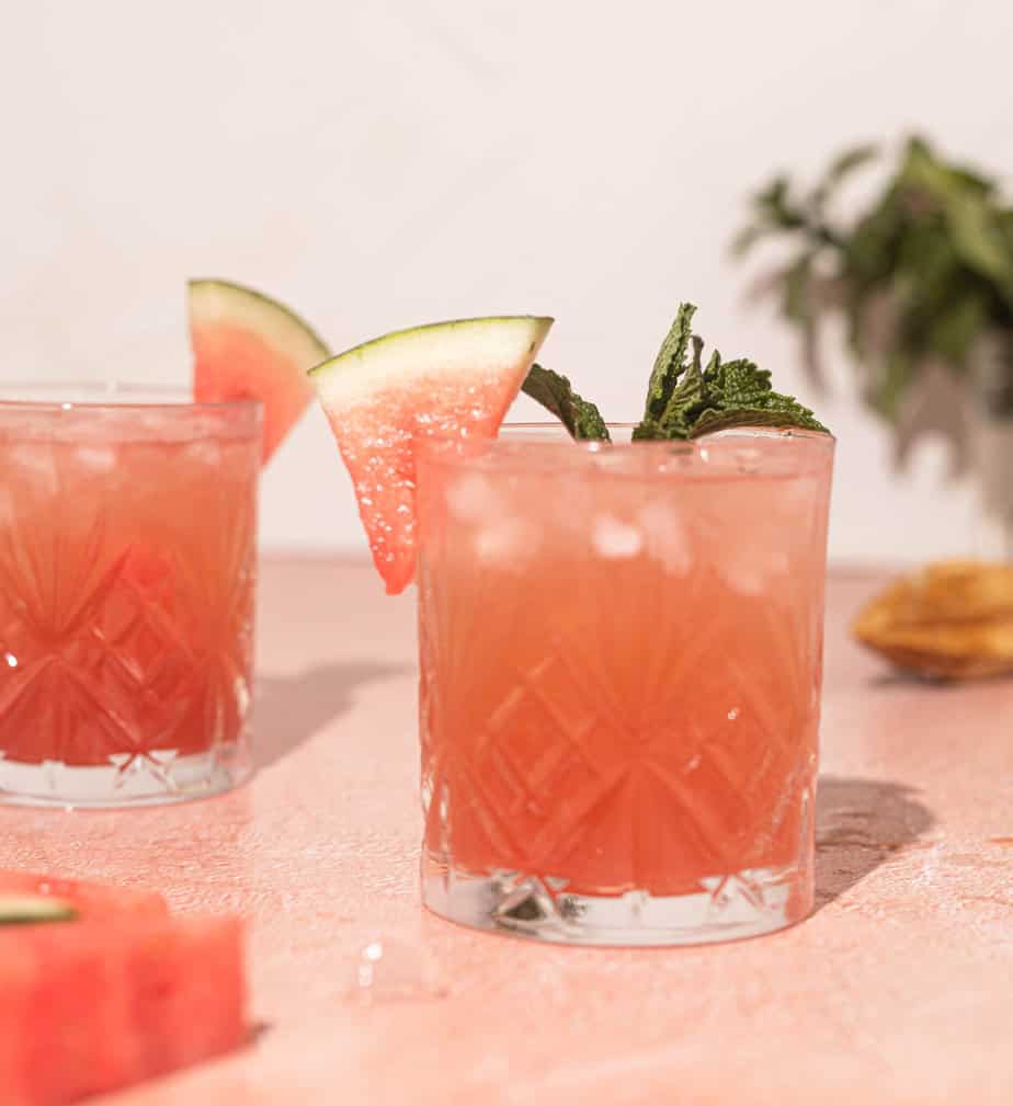 Watermelon Rum Cocktail in a glass with a watermelon slice and mint as a garnish on a pink surface with another cocktail in the background and a wooden juicer on the side 