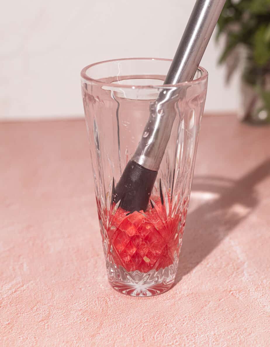 A shaker glass with watermelon and a muddler in the inside of the glass showing the juice released from the watermelon.