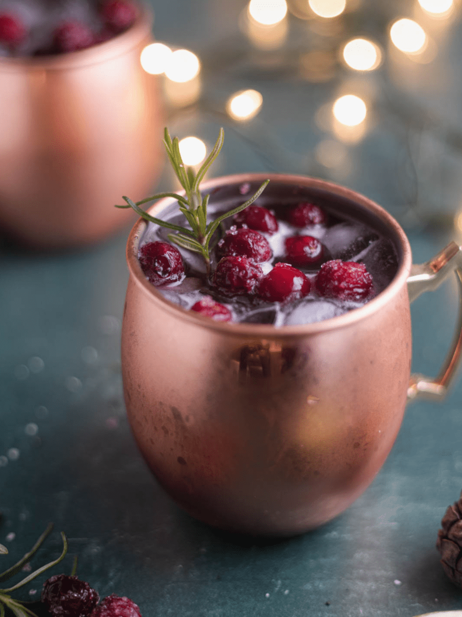 Christmas moscow mule in a copper mug with a sugared cranberry garnish and rosemary with twinkle lights in the background a