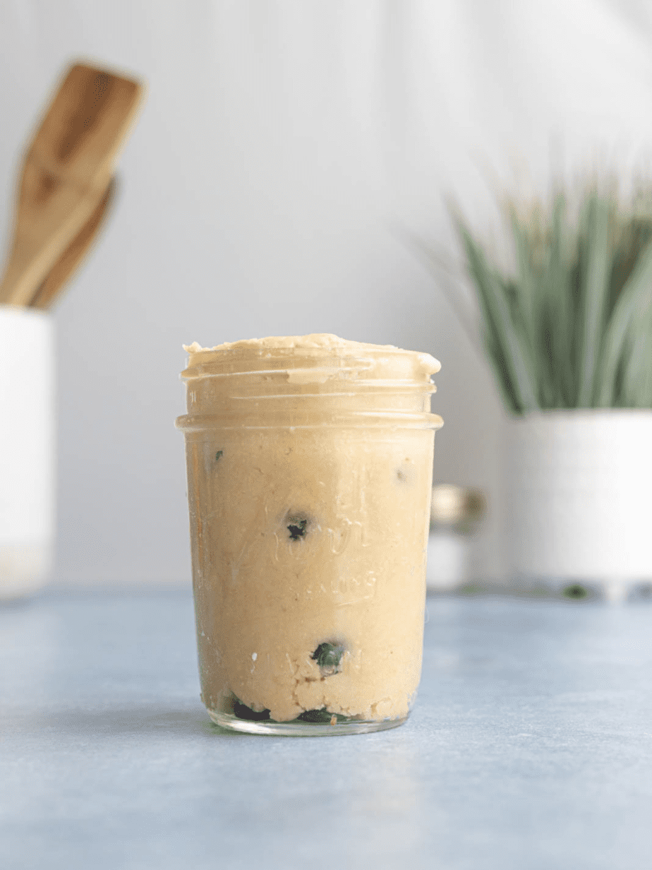 cookie dough in a mason jar on a blue surface with a plant and spoons in background
