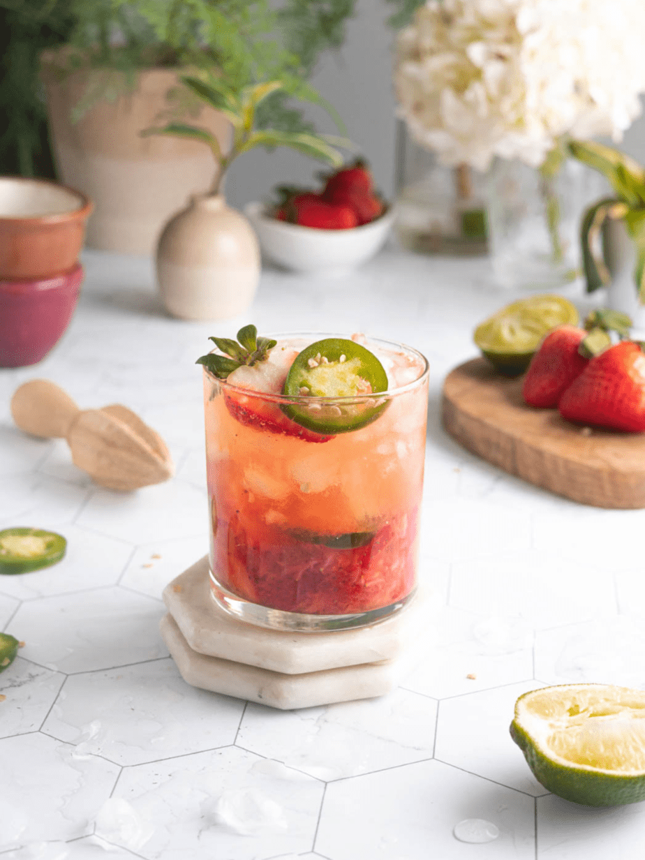 Jalapeno Strawberry Margarita in a glass with ice and a sliced strawberry and jalapeno on top with flowers in the background