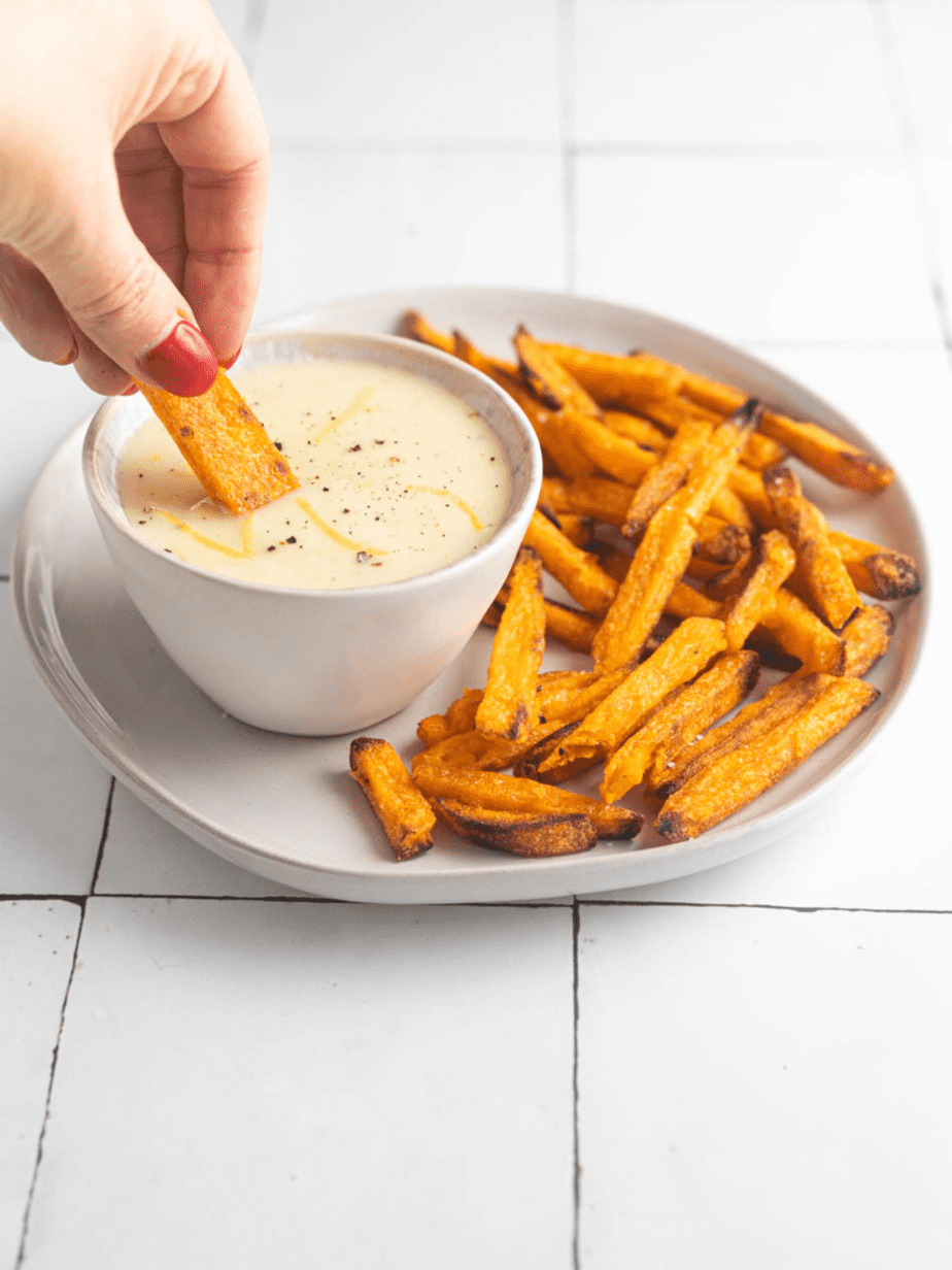 Vegan Garlic Aioli in a bowl on top of a plate with sweet potato fries and a fry being dipped into the aioli 