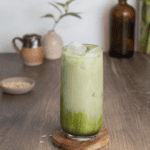 Iced Matcha Oat Milk latte on a wood surface with oats in front of it and plants in the back