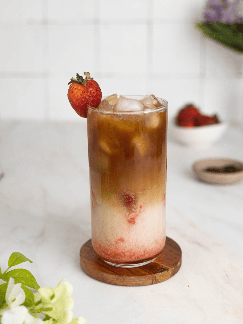 Iced strawberry latte not mixed (with layers for each ingredient) with a strawberry garnish with a bowl of coffee beans and a bowl of strawberries in back
