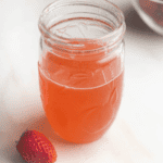 Strawberry simple syrup in a mason jar with strawberry next to it and strawberry bowl in background