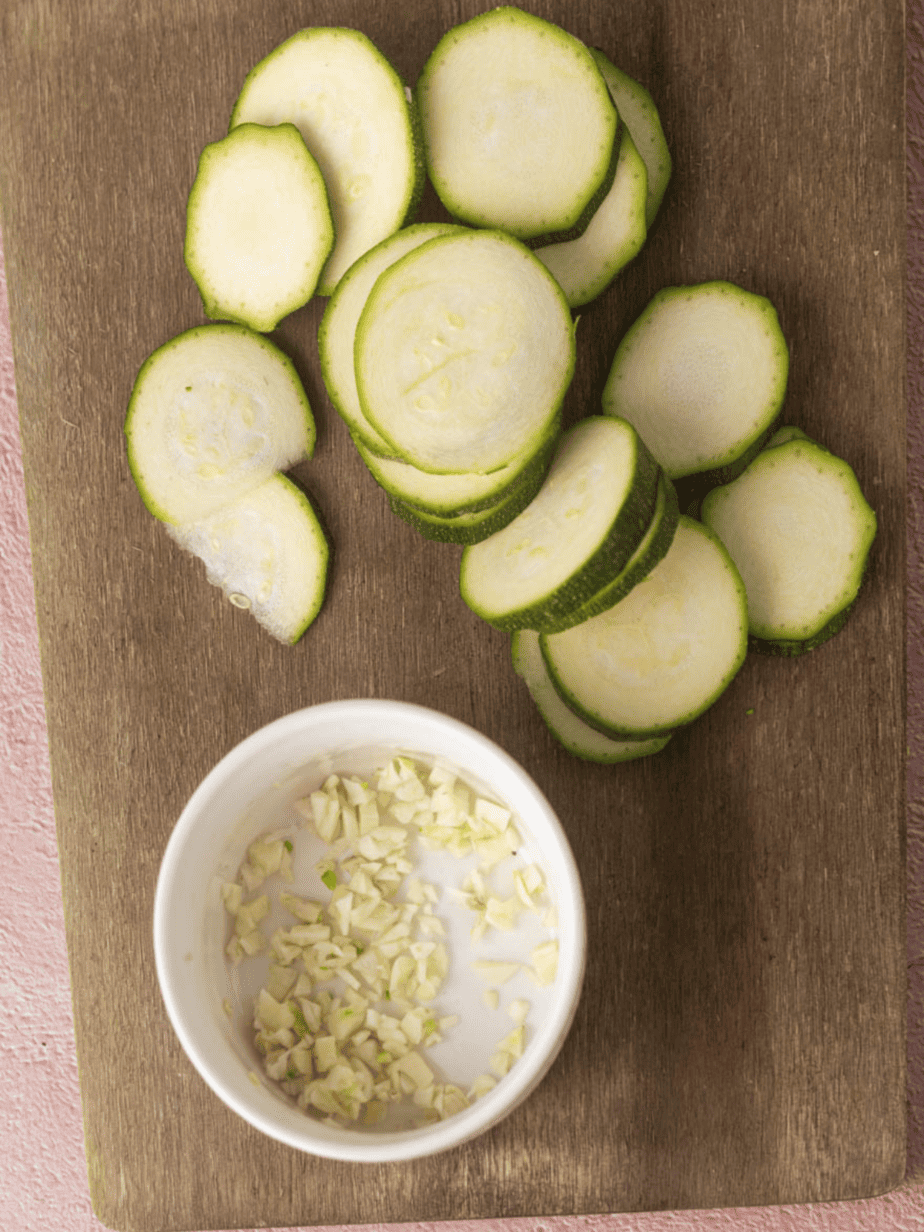 zuchinni sliced into thin slices on a cutting board with minced garlic in a bowl next to it