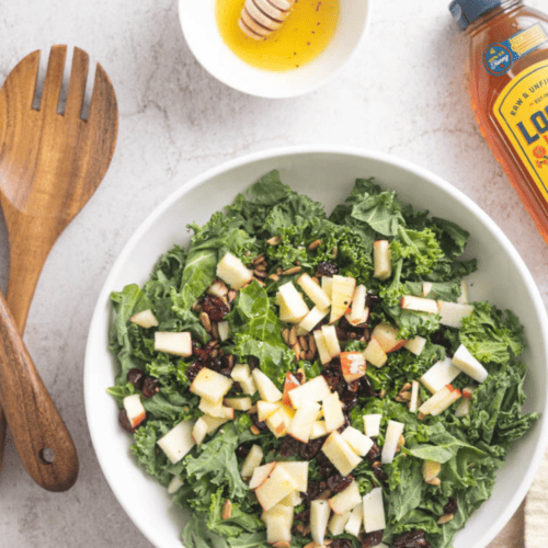 Kale and apple Manchego salad in a bowl with wood salad spoon and fork next to it and dressing in a bowl