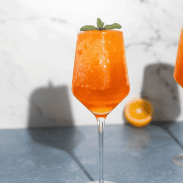 Frozen aperol spritz in glass with mint slice and orange behind it