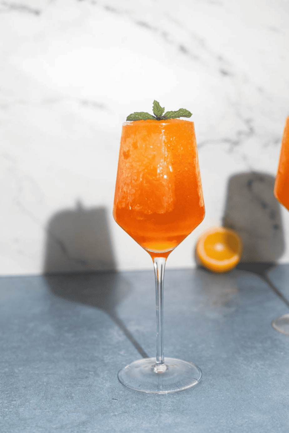 Frozen aperol spritz in glass with mint slice and orange behind it 